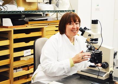Judith in the lab, sitting down looking into microscope and smiling into camera